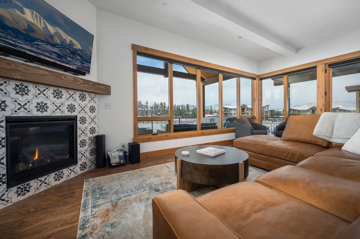 This interior photo centers on a welcoming living area with a prominent stone fireplace, a large TV, a leather couch, and expansive windows showcasing the natural surroundings. The atmosphere is warm and inviting, perfect for gathering, socializing, and enjoying cozy evenings.