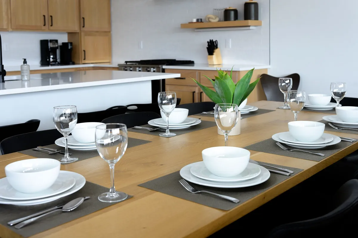 A stylish dining area centered around a table set with plates and glasses, complemented by a green plant centerpiece. The kitchen is visible in the background, reinforcing the open-plan layout and the connection between cooking and dining.