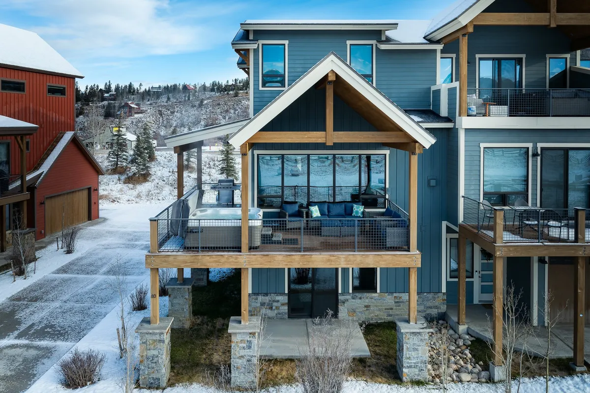 The image showcases the exterior of a luxurious mountain villa in Fraser, Colorado. Key features include a spacious deck with a private hot tub, large windows offering panoramic views, striking wooden beams, and a sturdy stone foundation. The scene evokes a sense of comfort and serenity, ideal for relaxing in the hot tub while watching snow fall, hosting summer barbecues, or stargazing from the deck with friends and family.